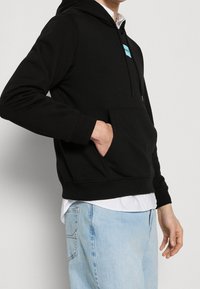 Sudadera negra con capucha hecha de tela suave con un bolsillo frontal y una etiqueta con logo azul, llevada sobre una camisa blanca y combinada con jeans azul claro.