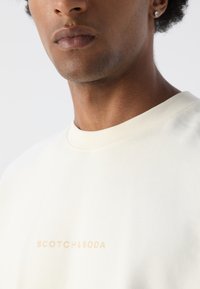 Creme farvet kortærmet t-shirt med rund halsudskæring, prydet med et diskret beige logo "SCOTCH & SODA" trykt på forsiden. Blød tekstur.