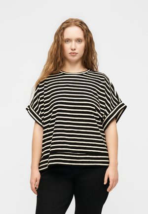 Junge Frau mit langen lockigen Haaren, die ein schwarz-weiß gestreiftes Kurzarmshirt und schwarze Hose trägt, vor einem einfarbigen Hintergrund stehend.