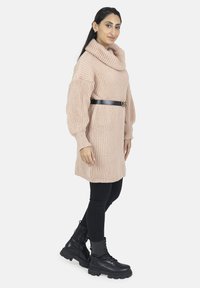 Vestido suéter oversized de punto beige con cuello de tortuga, textura acanalada y cinturón de cuero negro. Combinado con leggings negros y botas de combate.