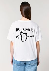 Een persoon draagt een oversized witte t-shirt met een grafisch ontwerp op de achterkant dat "Mi Amor" met een hart en pijlen toont.