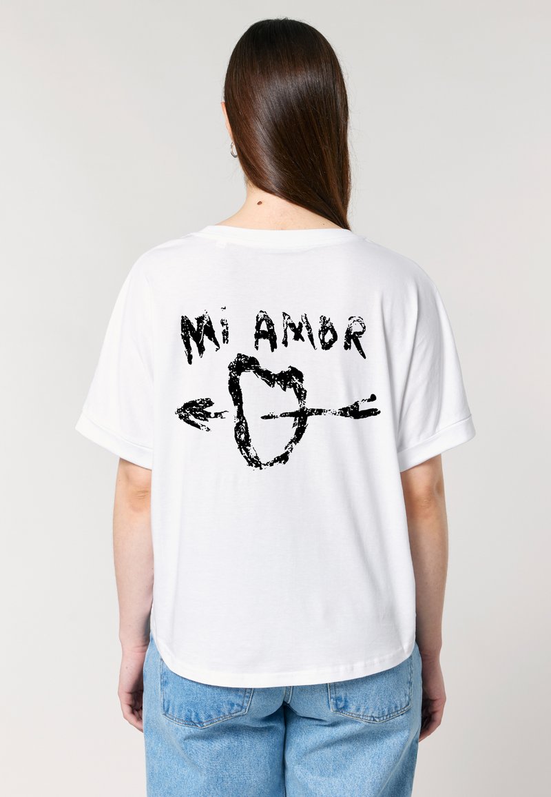 Een persoon draagt een oversized witte t-shirt met een grafisch ontwerp op de achterkant dat "Mi Amor" met een hart en pijlen toont.