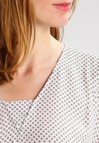 Bílá blůza s véčkovým výstřihem, zdobená geometrickým vzorem v béžové a hnědé barvě. Lehká tkanina, hladká textura a přiléhavý střih.