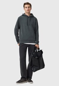 Felpa grigia con cappuccio e lacci, logo nero, abbinata a jeans scuri e sneakers grigie. Il modello tiene una borsa tote nera con dettagli del logo.