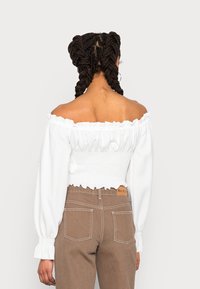 Vit off-shoulder topp med volangkant och smockad midja, kombinerad med bruna högmidjade jeans med kontrasterande sömmar och bakfickor.