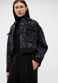 Fiatal nő, aki fekete és kék texturált rövid dzsekit visel fekete garbó fölött, fekete magas derekú nadrággal, egyik kezét a zsebében tartva pózol.