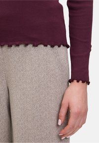 Haut à manches longues côtelé bordeaux avec un ourlet à volants, associé à un pantalon à motif pied-de-poule marron clair. Texture du tissu visible, main reposante.