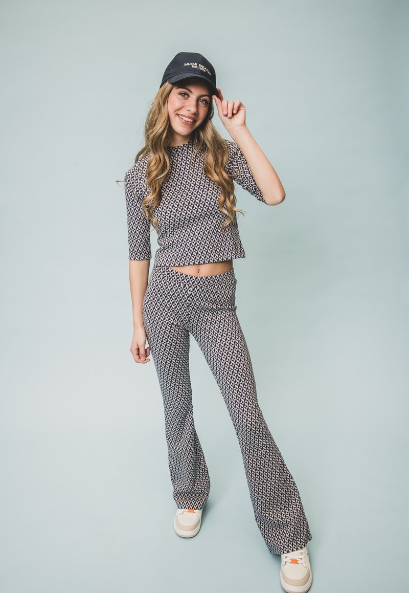 Twee-delige outfit bestaande uit een cropped top en flared broek van een marineblauwe stof met een geometrisch wit patroon. Gekleed met een zwarte pet en sneakers.