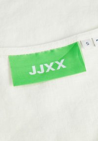 Hvidt stof med et grønt mærke, der viser "JJXX" i hvid skrift. Mærket angiver også størrelse "S" med ekstra mærker til stede.