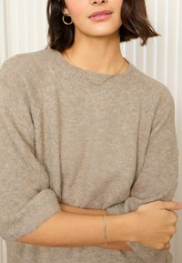 Beige Strickpullover mit lockerer Passform und aufgerollten Ärmeln, kombiniert mit goldenen Creolen und einem dünnen Kettenarmband am Handgelenk.