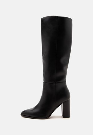 Botte en cuir noire montée jusqu'au genou avec bout pointu et talon haut carré, vue de profil sur un fond clair uni.