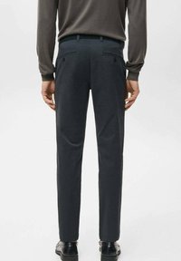 Mężczyzna w ciemnoszarych, dopasowanych spodniach garniturowych i czarnych eleganckich butach, stojący z rękami swobodnie opuszczonymi wzdłuż ciała na jednolitym tle.