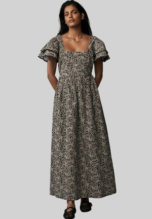 Frau trägt ein schwarzes Maxikleid mit kleinem weißem Blumenmuster, quadratischem Ausschnitt, gerafften kurzen Ärmeln und schwarzen flachen Schuhen.