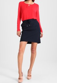 Blouse rouge à manches longues avec un décolleté en trou de serrure, associée à une jupe portefeuille bleu marine. Le mannequin porte des chaussures à talons bloques de couleur rose pâle. Fond uni.