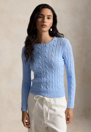 Γυναίκα με μακριά σκούρα μαλλιά που φοράει ένα ανοιχτό μπλε πουλόβερ πλεκτό με μικρό κίτρινο λογότυπο και λευκό παντελόνι με κορδόνι.