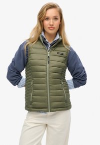 Olivgrön vadderad väst med dragkedja fram, sidofickor och hög krage. Bärs över en blå långärmad tröja med randig manschett och krämfärgade byxor.