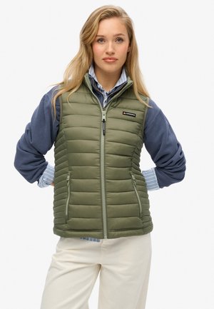 Gilet rembourré vert olive avec fermeture éclair sur le devant, poches latérales et col montant. Porté sur une chemise bleue à manches longues avec un poignet rayé, et un pantalon crème.