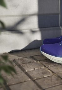 Coppia di scarpe da ginnastica viola con suole bianche appoggiate su un pavimento in mattoni testurizzati vicino a un muro di pietra.