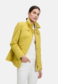 Gelbe leichte Jacke mit Stehkragen, Reißverschlusstaschen und lässiger Passform, kombiniert mit einem weißen Oberteil und hellen Hosen.