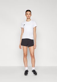 T-shirt de sport blanc à manches courtes avec un logo Adidas, associé à un short noir et des chaussures de sport noires. Fond uni.