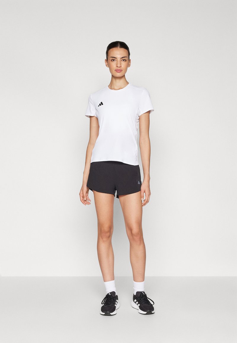 Camiseta atlética blanca de manga corta con el logo de Adidas, combinada con pantalones cortos negros y zapatillas deportivas negras. Fondo simple.
