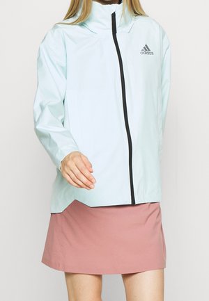 Femme portant une veste Adidas bleu clair avec une fermeture éclair noire et une jupe rose poussiéreuse, debout devant un fond clair uni.