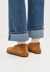 Bruine suède enkelboots met een rubberen zool, gedragen met opgerolde blauwe spijkerbroeken en lichtgekleurde geribbelde sokken. Eenvoudig ontwerp, geen opvallende patronen.