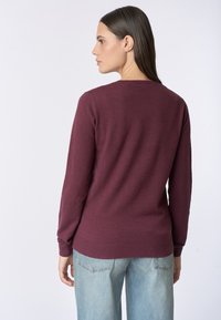 Maglione bordeaux con maniche lunghe e scollatura rotonda, realizzato in un tessuto morbido. L'orlo ha una texture a costine; indossato con jeans azzurri chiari.