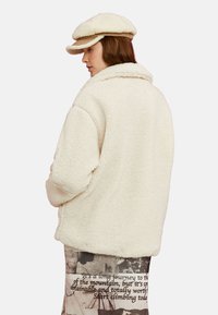 Chaqueta de textura cremosa y esponjosa con cuello y un corte oversize. Se lleva con una gorra color tostado, mostrando un estilo relajado y casual.