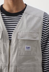 Gilet grigio chiaro realizzato in tessuto testurizzato, dotato di una zip frontale, due tasche a toppa e un logo Lee blu e bianco, indossato sopra una camicia a righe in bianco e nero.
