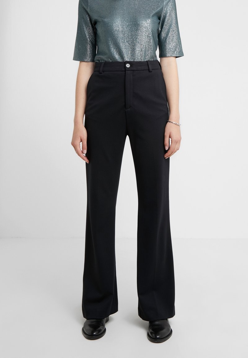 Femme portant un pantalon noir taille haute sur mesure avec bouton et fermeture éclair, associé à un haut métallique à manches courtes et des chaussures noires.