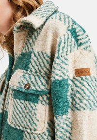 Fleece shirtjack in crème en teal met een geruit patroon. Beschikt over twee borstzakken en een leren logo patch. Gelaagde textuur.