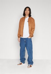 Jachetă bomber din suede maro, tricou alb, blugi albaștri și adidași cu model. Modelul stă în fața unui fundal simplu, cu mâinile în buzunare.
