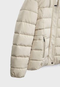 Beige pufferjakke med en quiltet design, der har en lynlåslomme og blød tekstur. Ærmerne har en rund form og tætsiddende pasform.