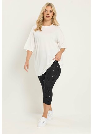 Frau geht, trägt ein übergroßes weißes T-Shirt, schwarze kurze Leggings mit weißen Punkten und weiße Sneakers vor hellem Hintergrund.