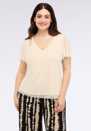 Femeie zâmbitoare cu părul închis la culoare, îmbrăcată cu o bluză crem cu mâneci transparente și pantaloni cu model tie-dye în nuanțe de negru și bej, stand în fața unui fond deschis.