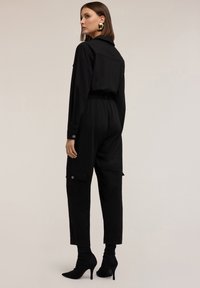 Zwarte jumpsuit met een cropped top, lange mouwen en elastische taille. Beschikt over cargozakken en is gestyled met zwarte enkelboots.