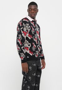 Zwart sweatshirt met opvallende grafische ontwerpen van handen en monden in rood en beige, met ribbelmanchetten en -zoom. Gecombineerd met een patroon van donkere broeken.