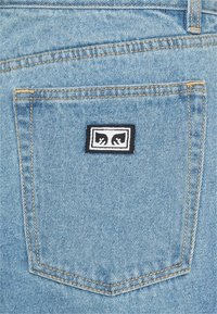 Lys blå denim med en baklomme. Har oransje sømmer og en rektangulær svart lapp med en hvit logo.