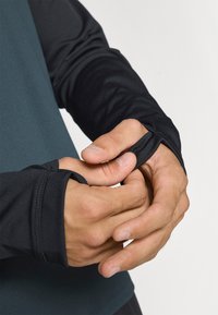 Zwart sportshirt met lange mouwen en duimopen, vervaardigd uit textuurrijk materiaal en met een contrasterend diepblauwe lijf. Close-up van handen die de mouwen aanpassen.
