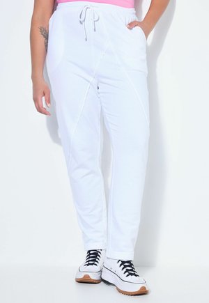 Persona che indossa pantaloni jogger bianchi con tasche frontali, una maglietta rosa e sneakers bianche con lacci neri, in piedi davanti a uno sfondo bianco.