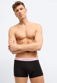 Μυώδης άνδρας με κοντά κατσαρά μαλλιά που φοράει μαύρα μποξεράκια Tommy Hilfiger με ροζ ζώνη στη μέση, στέκεται με σταυρωμένα τα χέ ρια μπροστά σε λευκό φόντο.