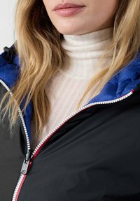 Giacca nera con cappuccio blu, zip in contrasto e fodera bianca. Indossata sopra un dolcevita crema con texture a costine e sottili righe.