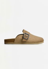 Beige suede-läpäisevä kenkä, jossa on metallisolki, korkki välipohja ja teksturoitu kumipohja.