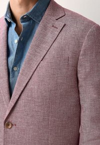 Blazer texturé rouge clair avec un revers à notch, fermeture à un bouton, et deux poches avant, superposé sur une chemise en denim bleue.