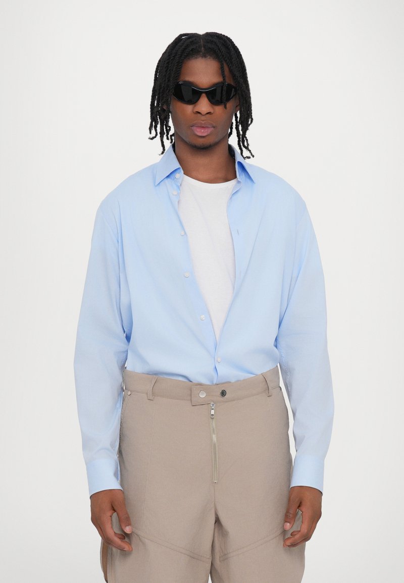 Light blue button-up shirt with a classic collar, paired with beige pants featuring a zippered front. Model wears black sunglasses.