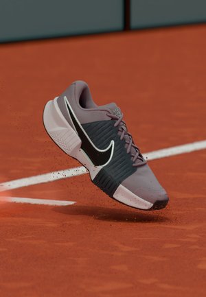 Chaussure de tennis Nike grise et noire avec semelle blanche soulevant de la poussière d'argile sur un court de tennis en terre battue orange près d'une ligne blanche.