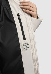 Veste doudoune de couleur claire avec un intérieur noir, dotée d'une poche zippée et d'une étiquette portant la mention : "Une étreinte chaleureuse faite de tissu."