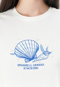 Hvid bomulds t-shirt med blå broderet muslingedesign, udsmykket med små perle accentuer. Teksten lyder "MUSLINGESØGER Siden 1996."