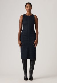 Γυναίκα με σκουρόχρωμα πλεκτά μαλλιά, ντυμένη με αμάνικο μαύρο τζιν φόρεμα μήκους μέχρι τη μέση της γάμπας και μαύρες δερμάτινες μπότες μέχρι το γόνατο, στέκεται μπροστά σε απλό φόντο.